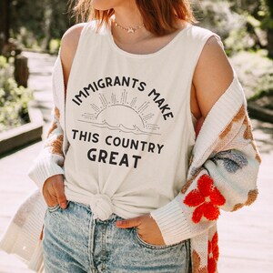 May include: Cream-colored tank top with the text "IMMIGRANTS MAKE THIS COUNTRY GREAT" in black. The design includes a sun and waves. The person is wearing a white and patterned cardigan and blue jeans.