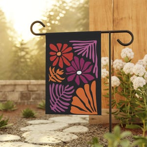 May include: A black garden flag featuring a floral design in shades of red, orange, and purple. The flowers and leaves are stylized with bold outlines. The flag is displayed on a black metal stand in a garden setting.
