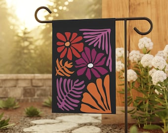 Op de afbeelding: Een zwarte tuin vlag met een bloemen design in rood, oranje en paarse tinten. De bloemen en bladeren zijn gestileerd. De vlag wordt getoond op een zwarte metalen standaard in een tuin.