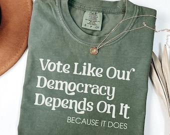May include: An olive green short-sleeve t-shirt with white text that reads "Vote Like Our Democracy Depends On It BECAUSE IT DOES". The shirt features a Comfort Colors brand label at the inner neckline and a delicate gold-toned chain necklace.