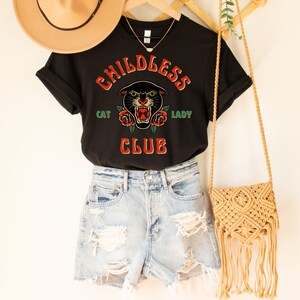 May include: Black t-shirt with a graphic of a black panther with red roses and the text "Childless Cat Lady Club". The shirt is being modeled with denim shorts and a straw bag.