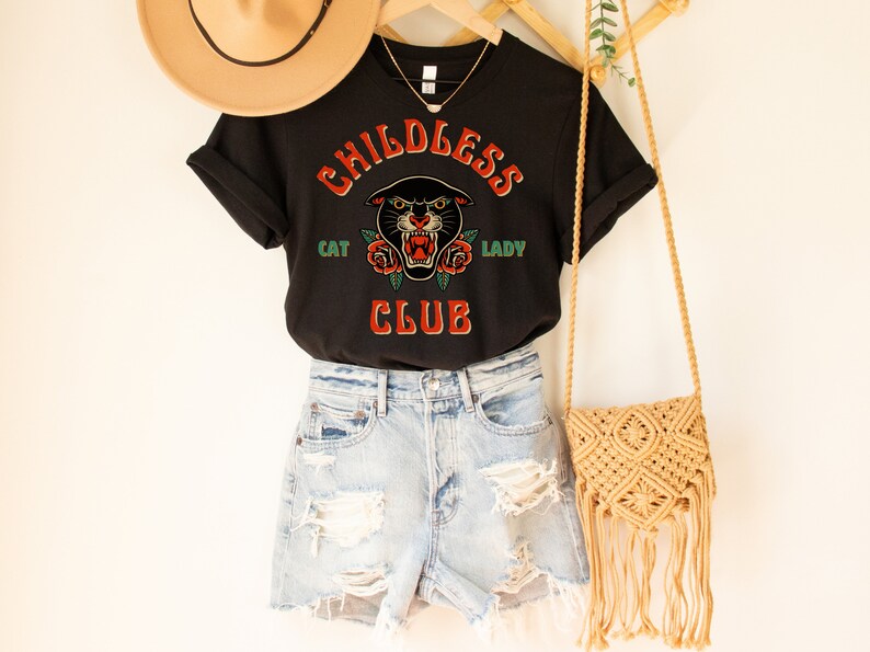 May include: Black t-shirt with a graphic of a black panther with red roses and the text "Childless Cat Lady Club". The shirt is being modeled with denim shorts and a straw bag.