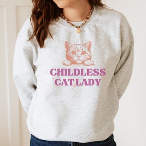 May include: A light gray sweatshirt with a pink graphic of a cat and the words "CHILDLESS CAT LADY" in pink. The cat is peeking over the text. The sweatshirt is a casual style.