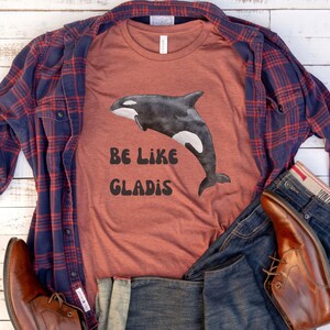May include: A rust-colored t-shirt with a black and white graphic of an orca whale and the text "Be Like Gladys". The shirt is styled with a blue and red plaid shirt, blue jeans, and brown leather boots.