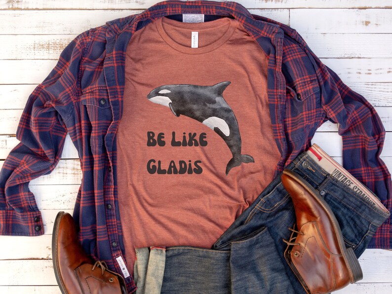 May include: A rust-colored t-shirt with a black and white graphic of an orca whale and the text "Be Like Gladys". The shirt is styled with a blue and red plaid shirt, blue jeans, and brown leather boots.