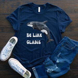 May include: A navy blue t-shirt with a watercolor illustration of an orca and the text "BE LIKE GLADIS" in white.