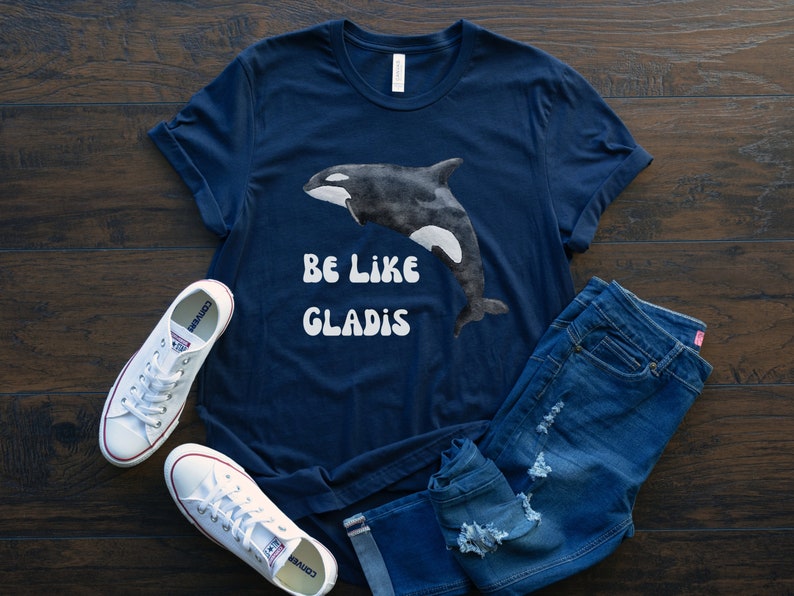 May include: A navy blue t-shirt with a watercolor illustration of an orca and the text "BE LIKE GLADIS" in white.