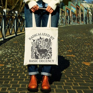 May include: Cream-colored tote bag with the words "RADICALIZED BY BASIC DECENCY" printed in a dark gray color. The bag features a graphic of a deer, rabbits, and foliage. The person is wearing blue jeans and brown boots.