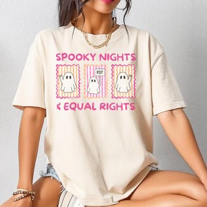May include: Cream-colored t-shirt with the text "SPOOKY NIGHTS & EQUAL RIGHTS" in pink. The design features three ghost illustrations within pink and orange frames. The middle ghost holds a sign that reads "FDT".