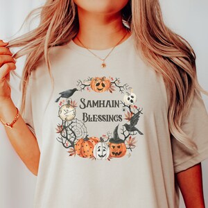 May include: A beige t-shirt with a watercolor design of a wreath with pumpkins, crows, a clock, and a spiderweb. The text "Samhain Blessings" is in the center of the wreath.
