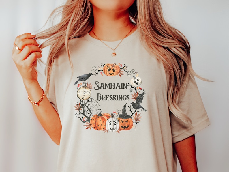 May include: A beige t-shirt with a watercolor design of a wreath with pumpkins, crows, a clock, and a spiderweb. The text "Samhain Blessings" is in the center of the wreath.