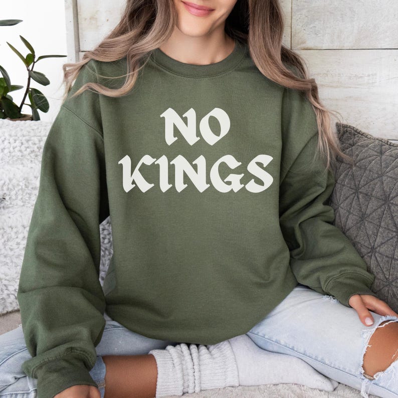 Puede incluir: Sudadera verde oliva con el texto "NO KINGS" en blanco. La sudadera es de cuello redondo y est&aacute; hecha de un material suave. La persona lleva vaqueros claros.