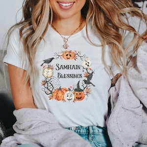 May include: White t-shirt with a graphic of a wreath of pumpkins, leaves, and a black bird. The text "Samhain Blessings" is in the center of the wreath.