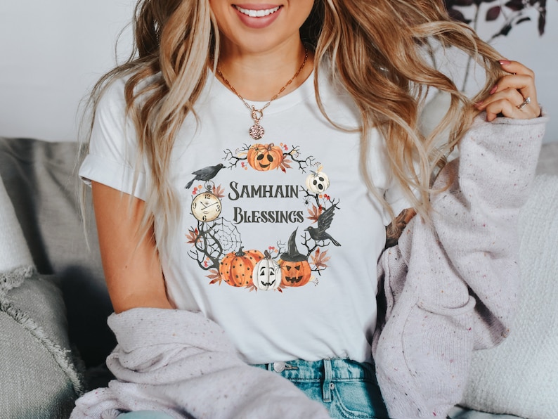 May include: White t-shirt with a graphic of a wreath of pumpkins, leaves, and a black bird. The text "Samhain Blessings" is in the center of the wreath.