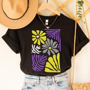 May include: Black t-shirt featuring a floral design with yellow, gray, white, and purple elements. The design includes stylized flowers and leaves. A tan hat and a gold necklace are also visible.