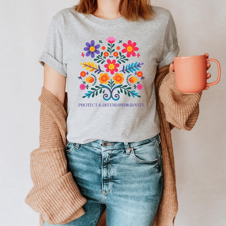 Puede incluir: Una camiseta gris con un dise&ntilde;o floral colorido y el texto "Proteger y defender a los inmigrantes".