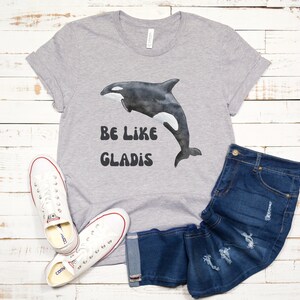 May include: Gray t-shirt with a black and white orca graphic and the text "Be Like Gladys" in a retro font.