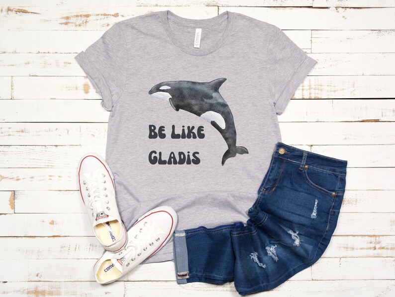 May include: Gray t-shirt with a black and white orca graphic and the text "Be Like Gladys" in a retro font.