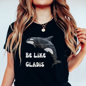 May include: Black t-shirt with a white orca graphic and the text "Be Like Gladys" in a retro font.
