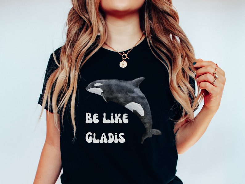 May include: Black t-shirt with a white orca graphic and the text "Be Like Gladys" in a retro font.