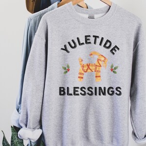 May include: A light gray sweatshirt with the words "YULETIDE BLESSINGS" in black. A straw goat illustration is centered above the word "BLESSINGS", with holly sprigs on either side. The sweatshirt is hanging on a wooden hanger.