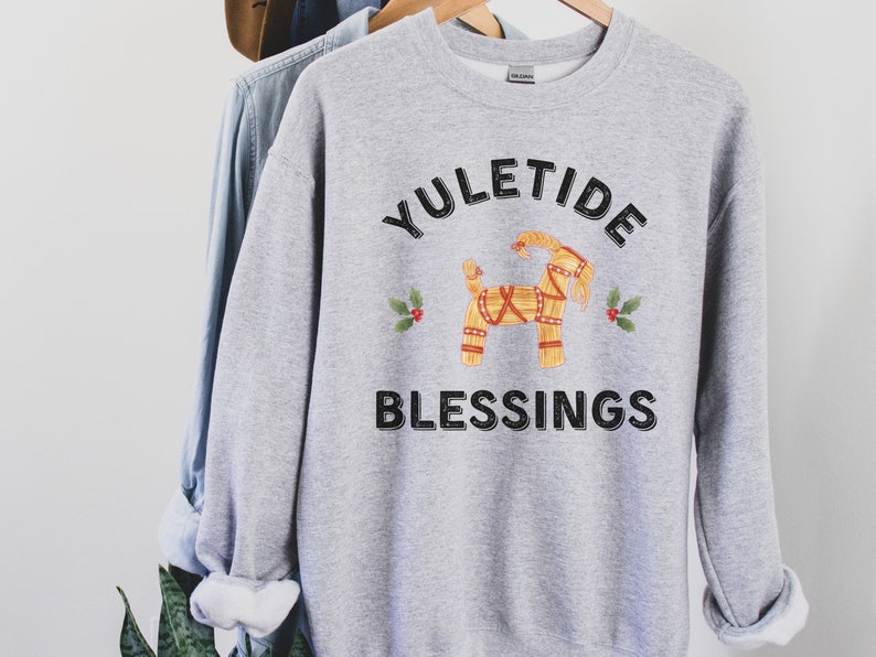 May include: A light gray sweatshirt with the words "YULETIDE BLESSINGS" in black. A straw goat illustration is centered above the word "BLESSINGS", with holly sprigs on either side. The sweatshirt is hanging on a wooden hanger.