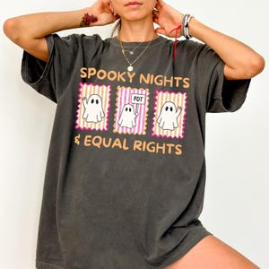 May include: Dark grey t-shirt with the text "SPOOKY NIGHTS & EQUAL RIGHTS". The shirt features three ghost illustrations inside pink and orange frames. One ghost holds a sign that reads "FDT".