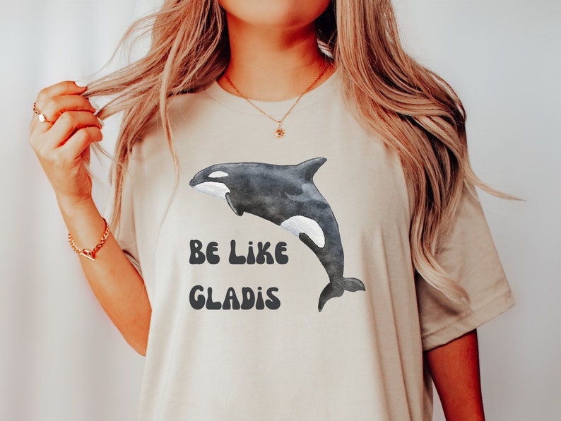 May include: A beige t-shirt with a black and white orca graphic and the text "BE LIKE GLADIS" in a retro font.