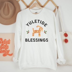 May include: White sweatshirt with the text "YULETIDE BLESSINGS" and a decorative Dala horse graphic. The design includes holly sprigs. A tan hat and a tote bag with red sunglasses are visible in the background.