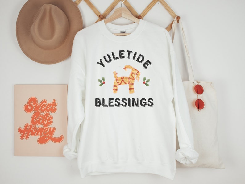 May include: White sweatshirt with the text "YULETIDE BLESSINGS" and a decorative Dala horse graphic. The design includes holly sprigs. A tan hat and a tote bag with red sunglasses are visible in the background.