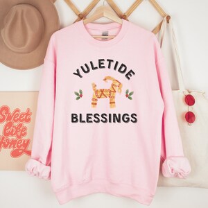 May include: A light pink sweatshirt with the words "YULETIDE BLESSINGS" in black. The design includes a straw goat and holly sprigs. The sweatshirt is hanging on a wooden hanger.