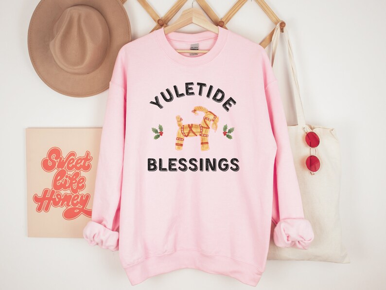 May include: A light pink sweatshirt with the words "YULETIDE BLESSINGS" in black. The design includes a straw goat and holly sprigs. The sweatshirt is hanging on a wooden hanger.