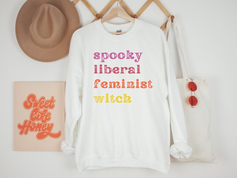 May include: White sweatshirt with the text "spooky liberal feminist witch" in rainbow colors.