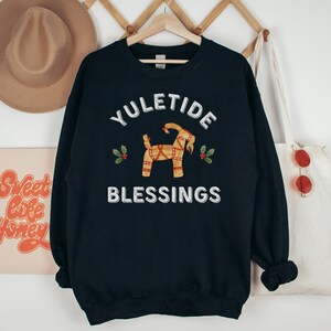 May include: A navy blue sweatshirt with the words "YULETIDE BLESSINGS" in white. The design includes a straw goat and holly sprigs. The sweatshirt is hanging on a wooden hanger.