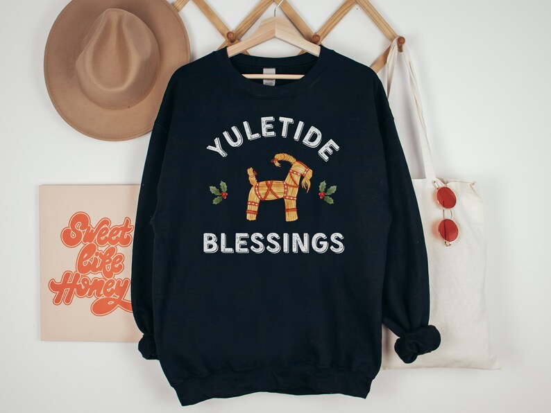 May include: A navy blue sweatshirt with the words "YULETIDE BLESSINGS" in white. The design includes a straw goat and holly sprigs. The sweatshirt is hanging on a wooden hanger.