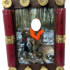 May include: A rustic photo frame with a hunting theme. The frame is made of dark brown wood and decorated with brass shotgun shell casings and red shotgun shells. The photo inside shows a hunter and a deer in a snowy forest.