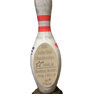 May include: A white bowling pin trophy with a brown wooden base. The pin has a gold plaque with the text "2024 Junior Gold Championships Detroit, MI Bobby Smith Avg - 201.6 64th Place".