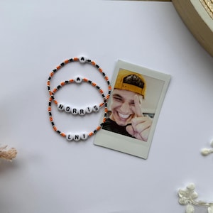 Peut inclure: Deux bracelets de perles avec les noms "NORRIS" et "LNI" épelés en perles de lettres blanches. Les bracelets sont noirs, orange et blancs. Une photo d'une personne souriante portant une casquette jaune est à côté des bracelets.