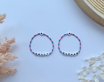 May include: Two beaded bracelets with pink and blue beads. The bracelets spell out the names "Ghastly" and "Doohan".