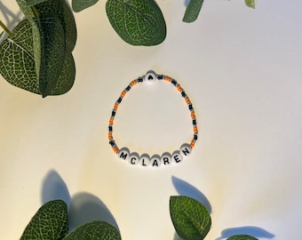 Peut inclure: Un bracelet de perles avec le nom "MCLAREN" épelé en perles de lettres blanches. Le bracelet comporte des perles orange, noires et argentées. Le bracelet est posé sur une surface blanche avec des feuilles vertes.