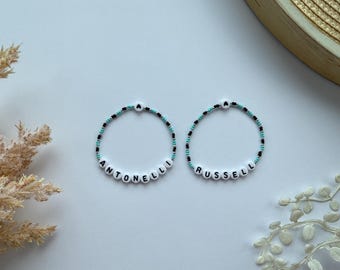 Puede incluir: Dos pulseras de cuentas con los nombres "ANTONELLI" y "RUSSELL" deletreados en cuentas blancas con letras negras. Las cuentas alternan negro y turquesa.