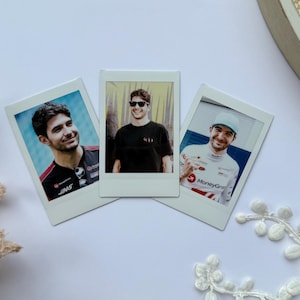 Op de afbeelding: Drie instantfoto's met een man in verschillende outfits. De foto's zijn gerangschikt op een wit oppervlak, met een decoratief bloemelement en een strohoed op de achtergrond. De man draagt op één foto een zonnebril.