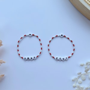 Op de afbeelding: Twee witte kralenarmbanden met rode kralen en zwarte letterkralen die "OB81" en "E031" spellen.