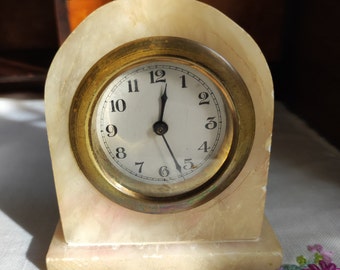 Könnte beinhalten: Eine kleine Vintage-Uhr mit einem cremefarbenen, marmorähnlichen Gehäuse und einer goldfarbenen Lünette. Das Zifferblatt ist weiß mit schwarzen Ziffern und Zeigern. Die Uhr ist rechteckig mit einem gewölbten Oberteil und steht auf einem kleinen Sockel.