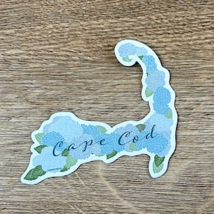 May include: A sticker in the shape of Cape Cod, Massachusetts, filled with blue and light blue hydrangea flowers. The words "Cape Cod" are written in cursive across the center of the sticker.