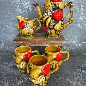 Pode incluir: Uma chaleira, um cremer e quatro canecas de cerâmica vintage decoradas com um motivo de frutas. A chaleira, o cremer e as canecas são todas marrons com detalhes verdes e vermelhos. O motivo de frutas inclui maçãs, peras, bananas e morangos.