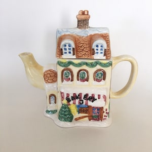 May include: A ceramic teapot shaped like a bakery building with a white snowy roof, brown brick chimney, and a green awning that reads "BAKERY". The teapot has a yellow base and brown details.