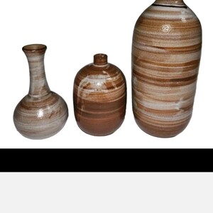 May include: A set of three ceramic vases featuring a horizontal banded pattern in shades of brown, tan, and white. The collection includes one tall slender vase, one medium-sized rounded vase with a narrow neck, and one shorter, wider vase.