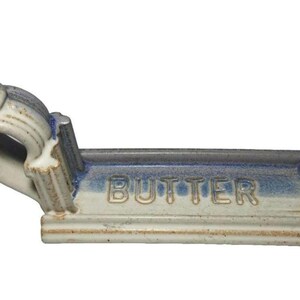 May include: A ceramic butter dish with a blue and white glaze. The word "BUTTER" is embossed on the front. The dish has a curved handle on one end and decorative detailing.