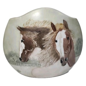 May include: A rounded ceramic vase featuring a hand-painted scene of two horses with brown and white coats. The horses are positioned side-by-side with detailed manes, set against a soft, muted background depicting a faint fence and landscape.